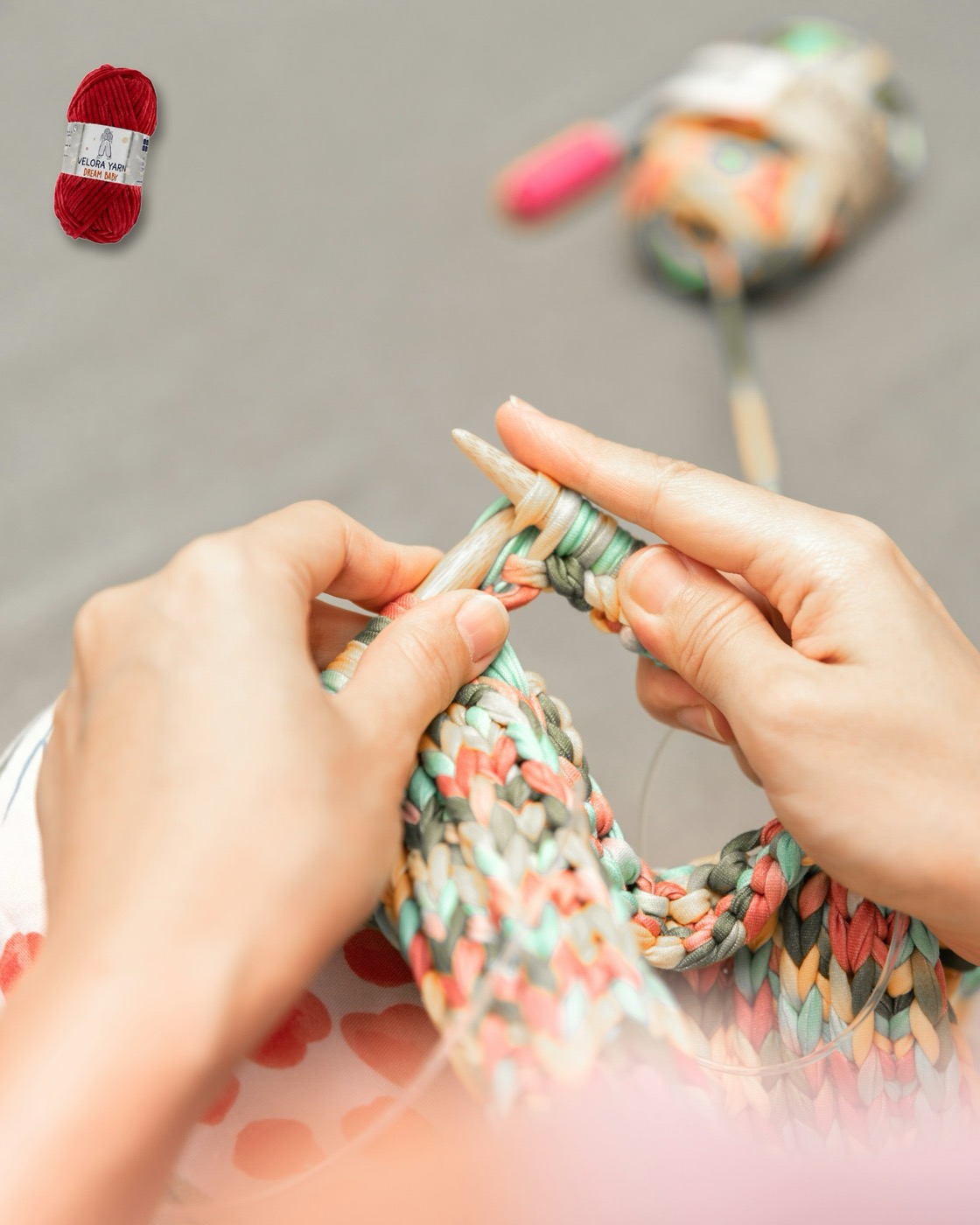 Hands knitting with thick chunky yarn on large needles, a skein of Velora Dream Baby chenille yarn nearby