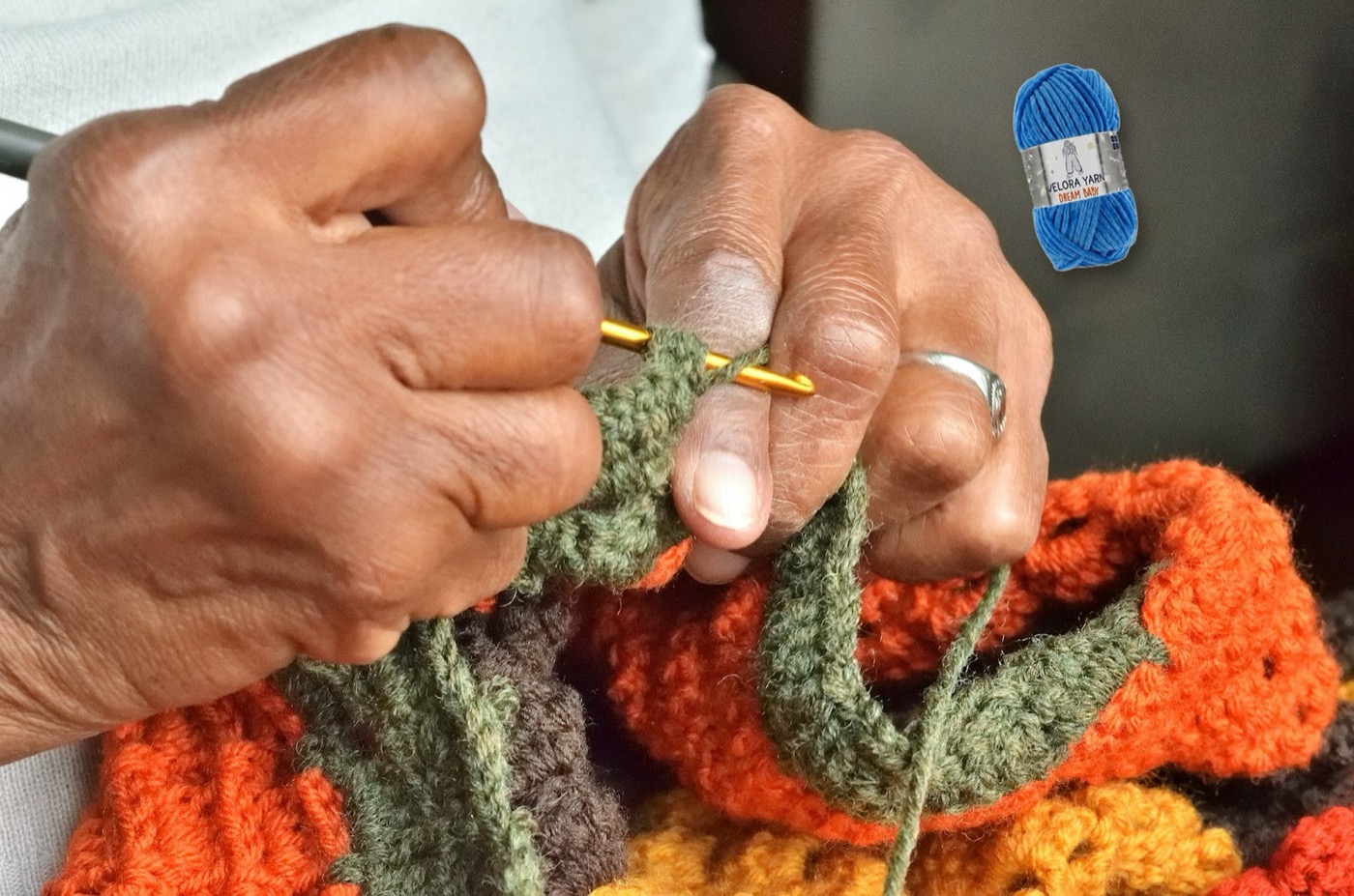Hands crocheting colourful granny squares, with a skein of Velora Dream Baby chenille yarn nearby