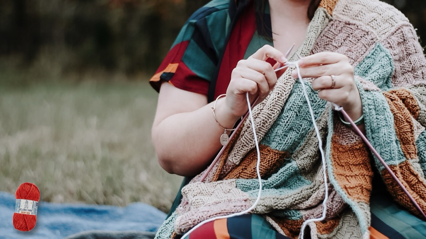 Woman knitting a colourful chunky blanket outdoors, with a skein of Velora Dream Baby chenille yarn beside her